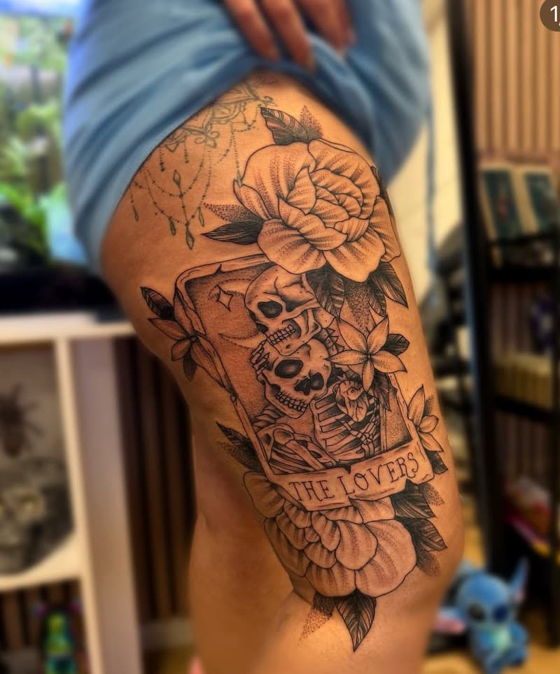 Blackwork tattoo van 'The Lovers' tarotkaart met skeletten en bloemen op bovenbeen.