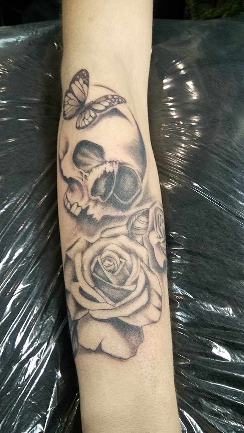 Zwart-wit tattoo van een schedel, roos en vlinder op de onderarm.