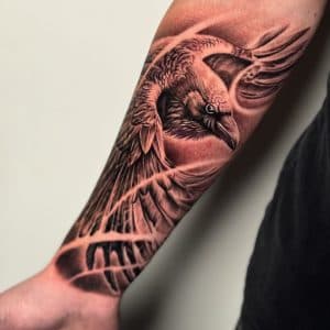 Realistische raaf tattoo op onderarm met vleugels en abstracte achtergrond.