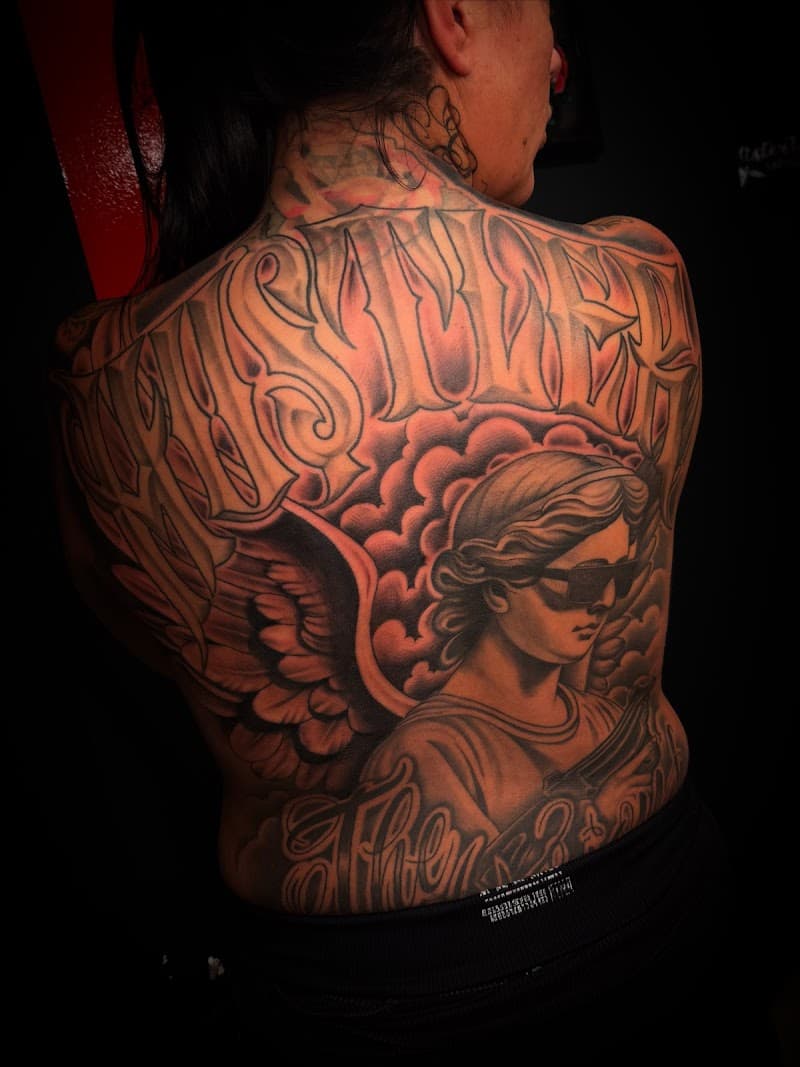 Grote zwart-wit rugtattoo met engel, wolken en tekst in Chicano stijl.