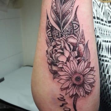 Zwart-wit tattoo met veer, bloemen en naam 'Nora' op de onderarm.