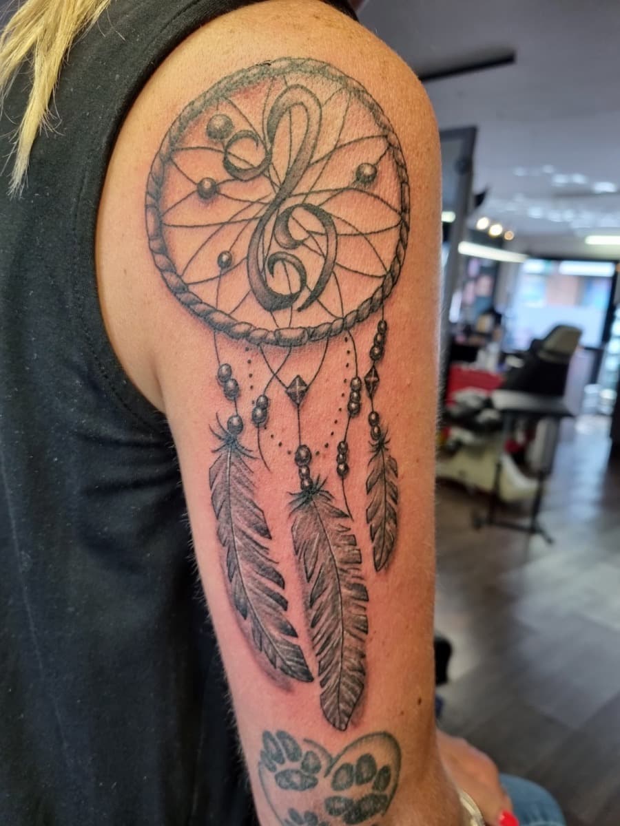 Zwart-grijze dromenvanger met muzieksleutel en veren op bovenarm.