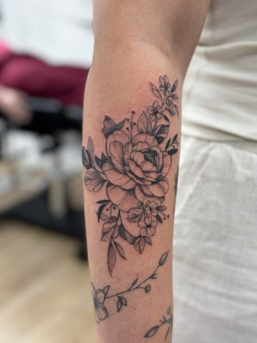 Fineline bloemen tattoo met pioenroos en bladeren op de onderarm.