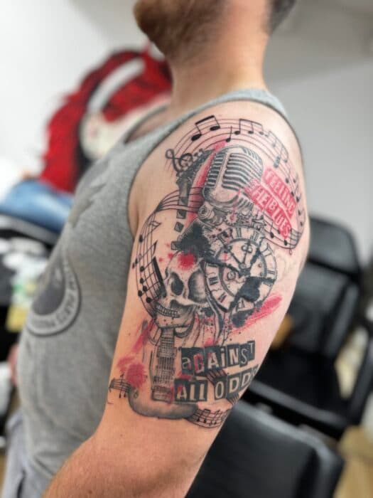 Trash polka tattoo met schedel, microfoon, klok en tekst op bovenarm.