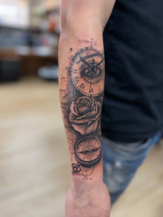 Realistische tattoo van klok, oog, roos en kompas op onderarm.