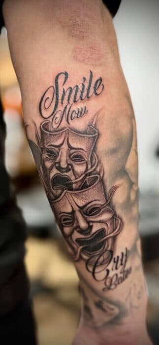 Smile Now Cry Later tattoo met theatermaskers en tekst op onderarm.