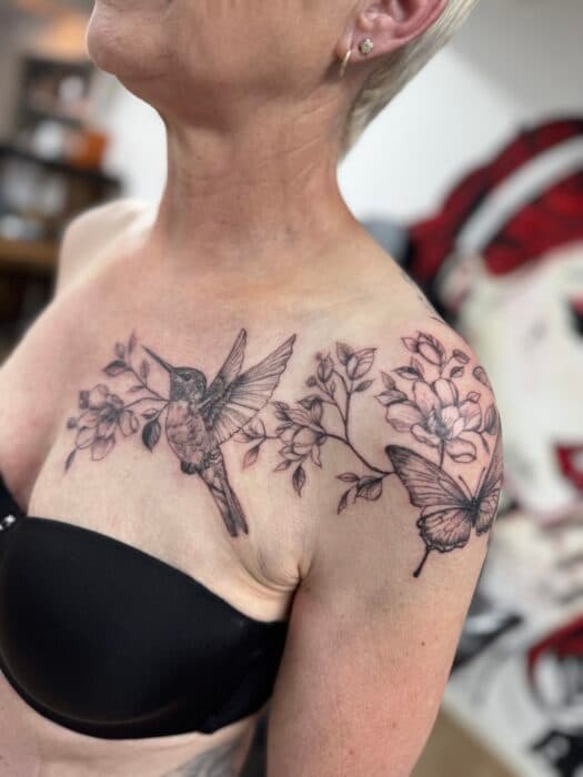 Fineline tattoo van kolibrie, vlinder en bloemen op borst en schouder.
