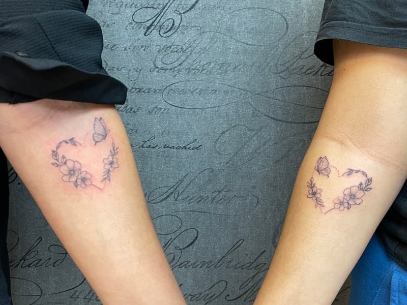 Twee identieke fineline tattoos van een hart gevormd door bloemen en een vlinder.