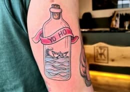 Kleurrijke tattoo van een fles met een papieren bootje en 'YO HO' banier.