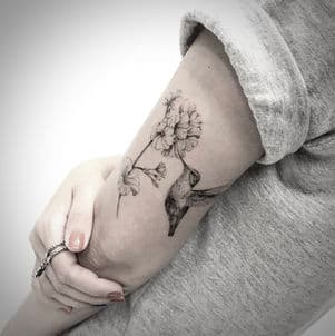 Fineline tattoo van een kolibrie bij bloemen op de bovenarm.