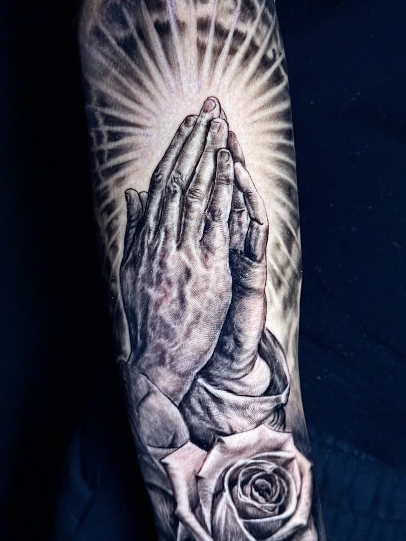 Realistische biddende handen met roos en stralen op de bovenarm.