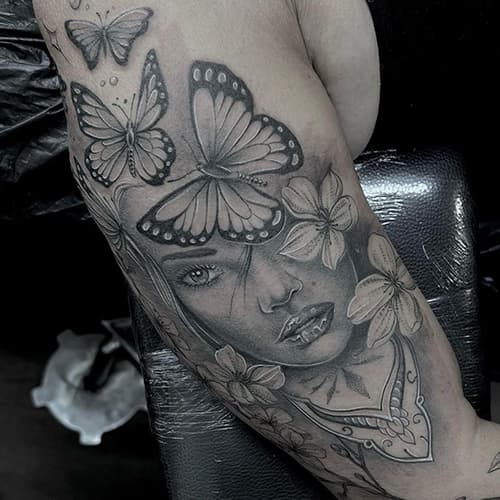 Black and grey tattoo van een vrouwengezicht met vlinders en bloemen op de bovenarm.