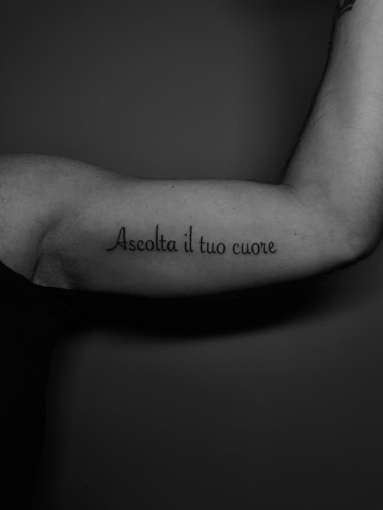 Fineline tekst tattoo 'Ascolta il tuo cuore' op de binnenkant van de bovenarm.