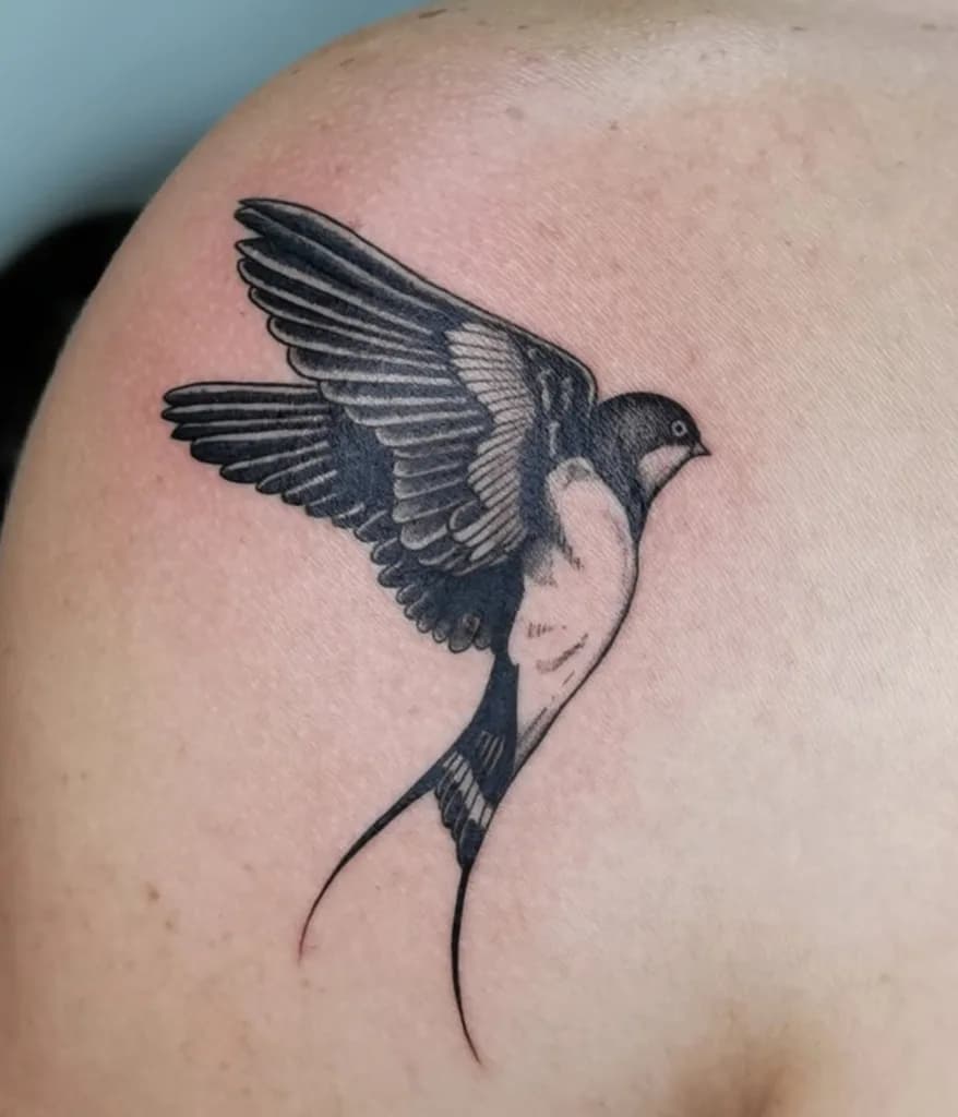 Zwart-grijze tattoo van een vliegende zwaluw op een schouder.