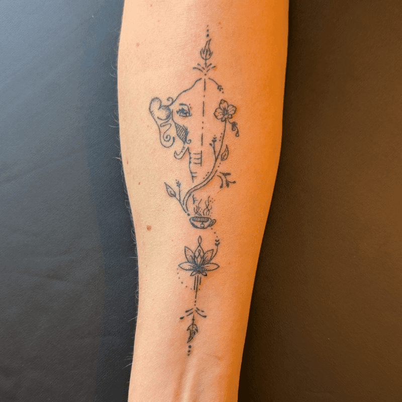 Fineline tattoo van Ganesha, lotus, bloemen en een kopje op de onderarm.