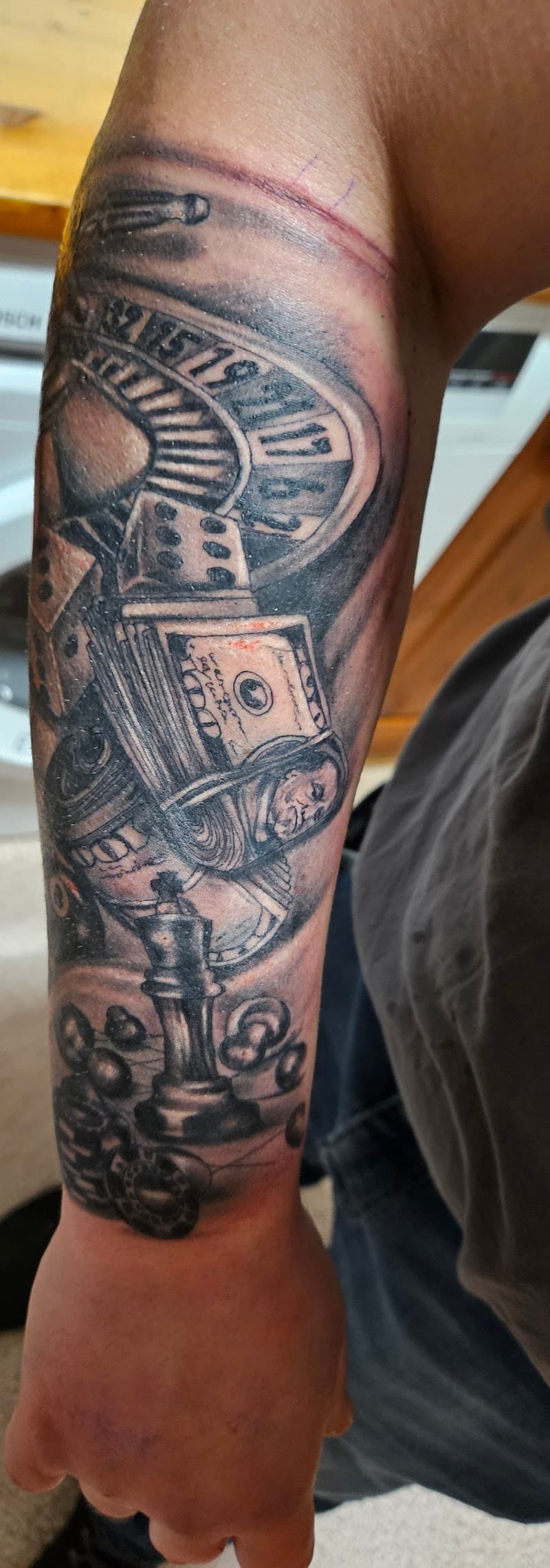 Zwart-wit tattoo met roulette, dobbelstenen, geld en schaakstukken op onderarm.