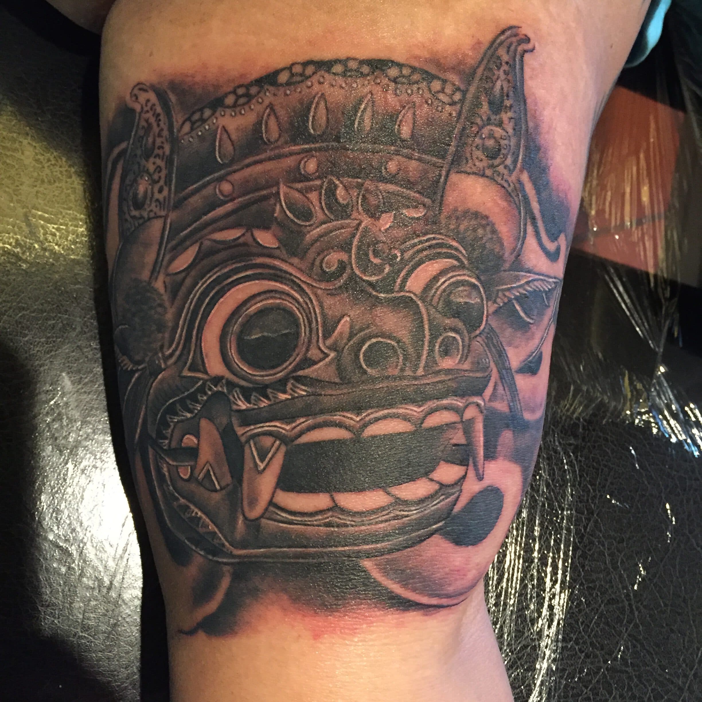 Zwart-wit tattoo van een Balinees Barong masker op de bovenarm.
