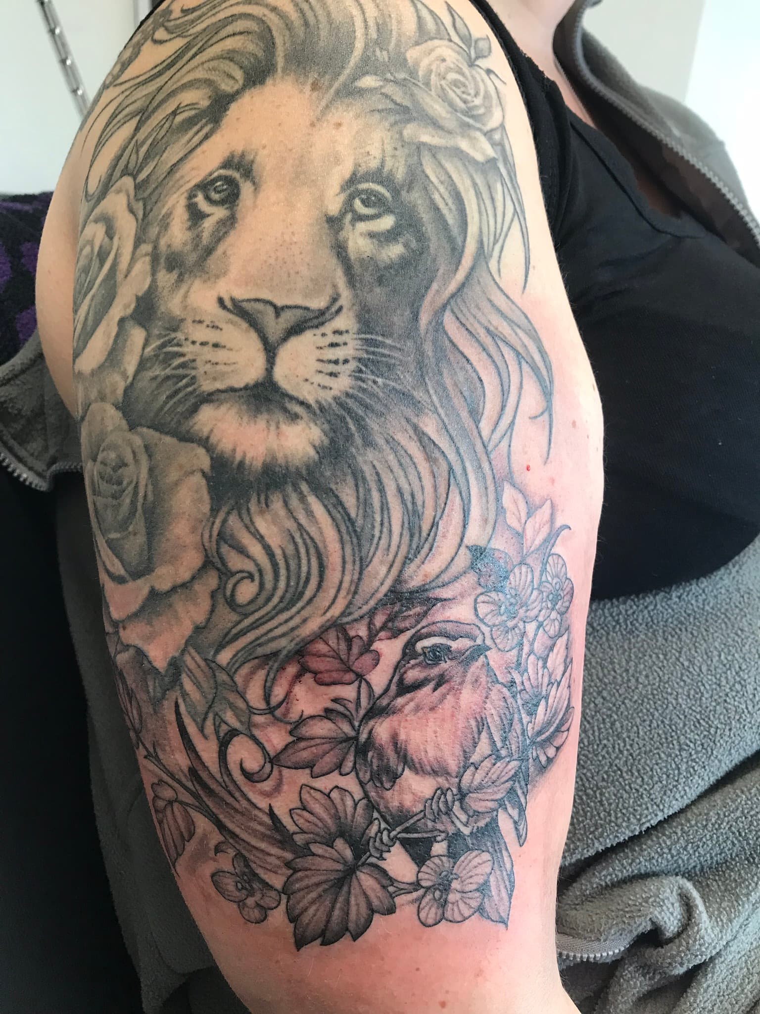 Grote zwart-wit tattoo van een leeuw, rozen en een vogel op de bovenarm.