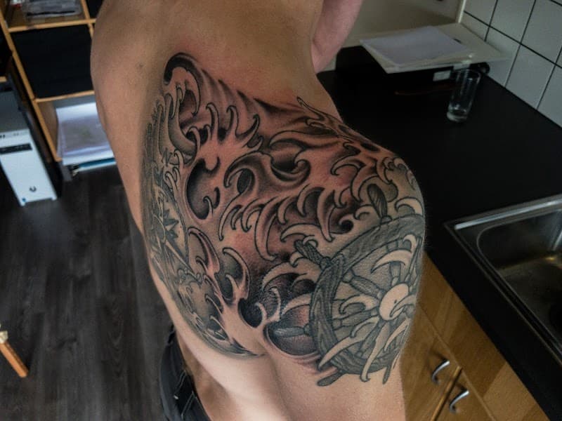 Zwart-wit tattoo van Japanse golven en een stuurwiel op de schouder.