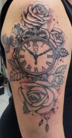 Zwart-wit tattoo van een zakhorloge met rozen en een vlinder op de bovenarm.