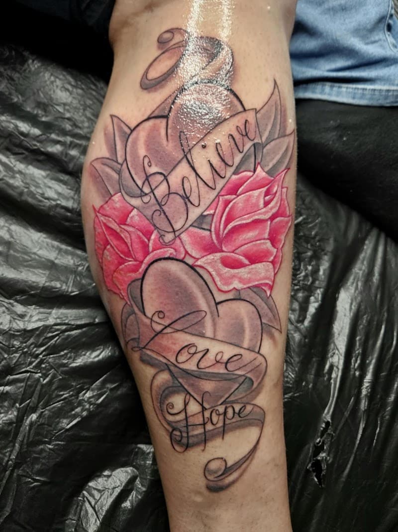 Neo-traditionele tattoo met harten, roze rozen en tekst 'Believe Love Hope' op onderbeen.