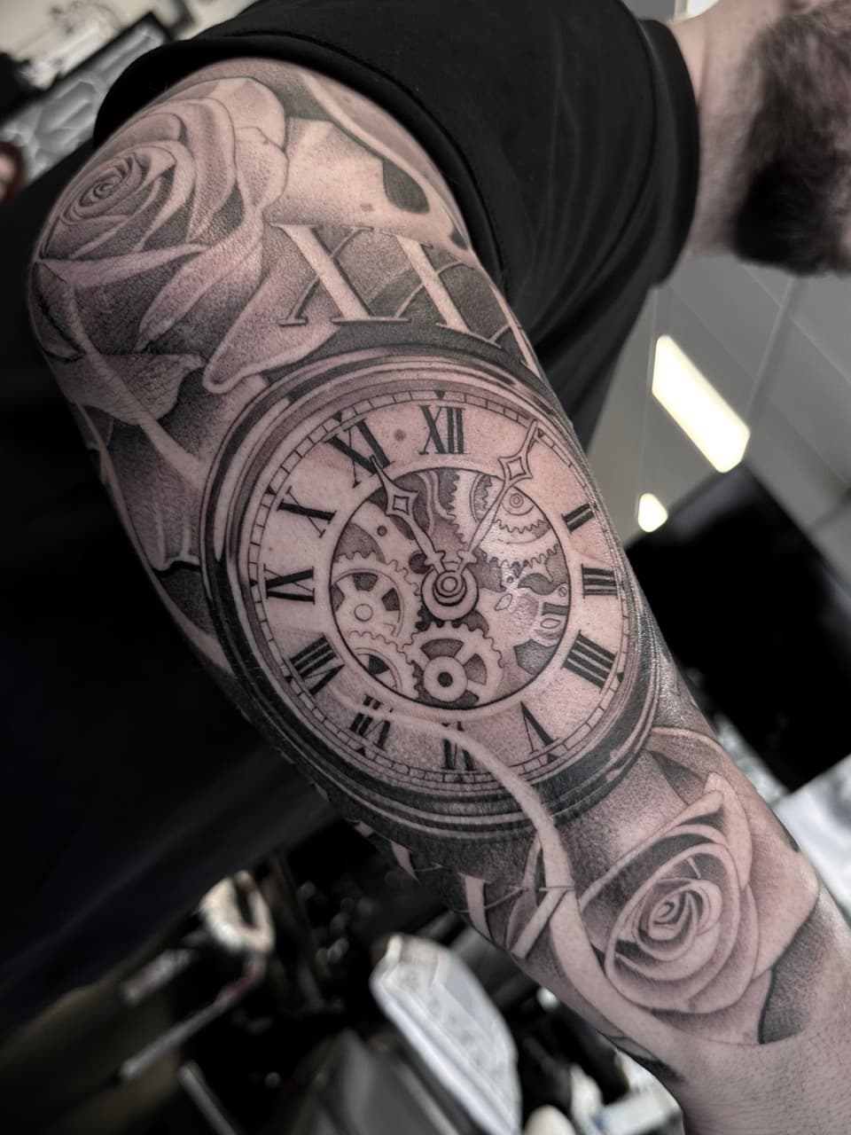 Zwart-wit tattoo van een zakhorloge met Romeinse cijfers en rozen op de bovenarm.