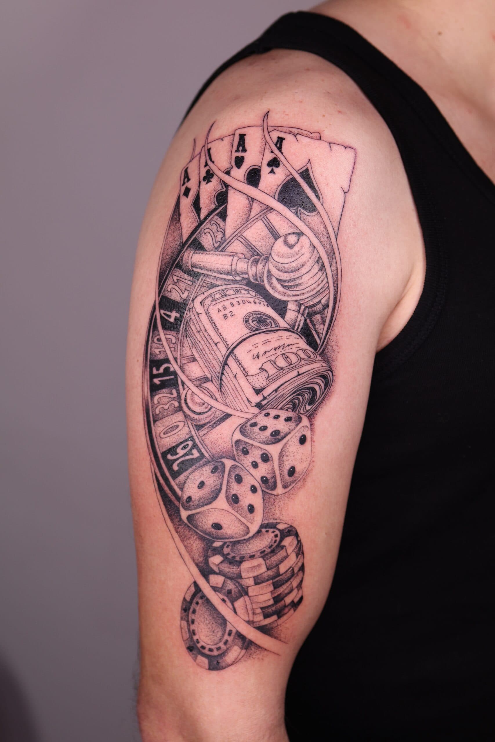 Zwart-wit tattoo met kaarten, dobbelstenen, geld en roulette op bovenarm.