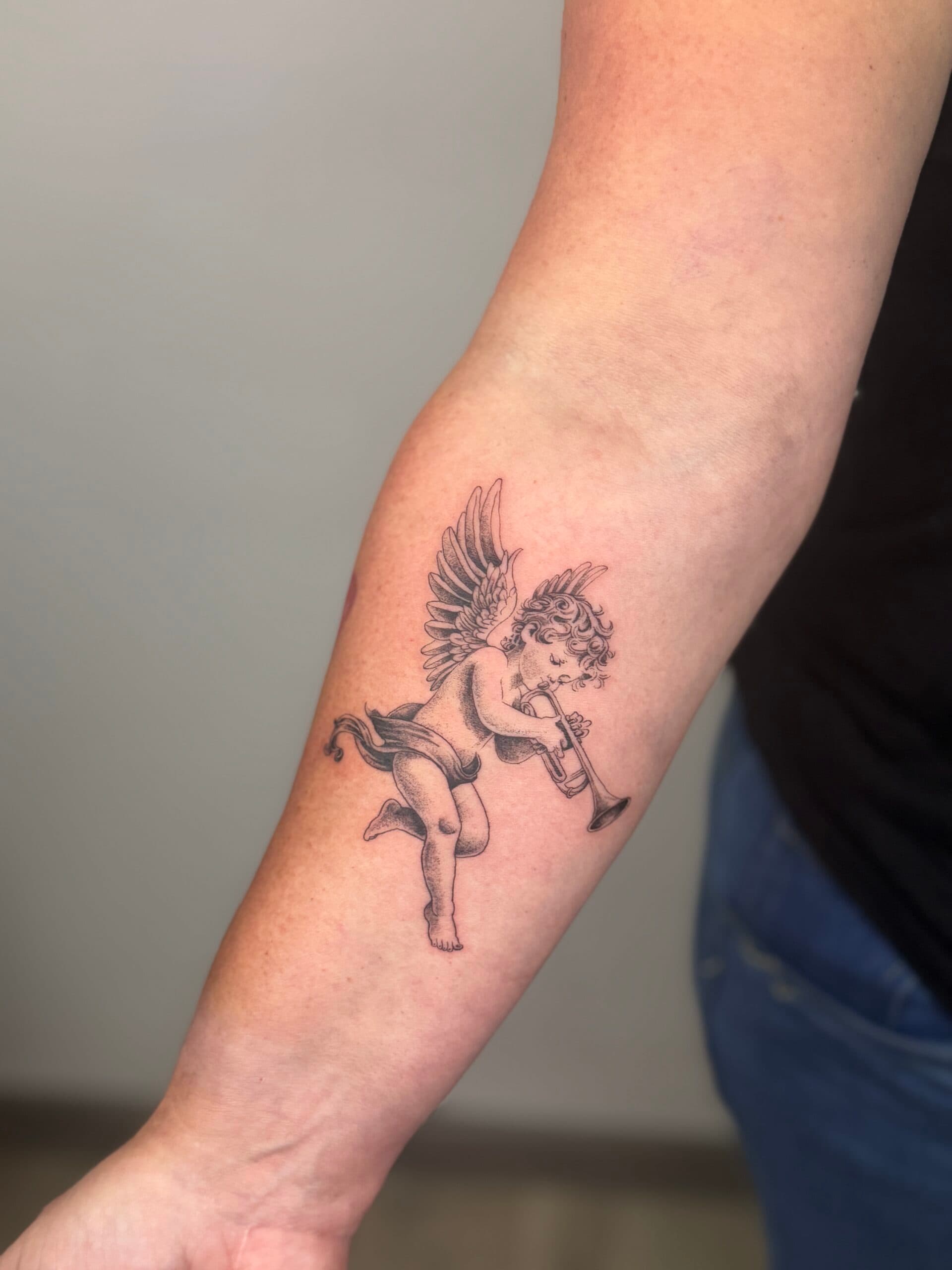 Fineline tattoo van een cherubijn met trompet op de onderarm.