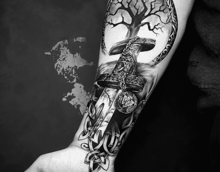 Zwart-wit tattoo van een zwaard, levensboom en Keltische knopen op de onderarm.