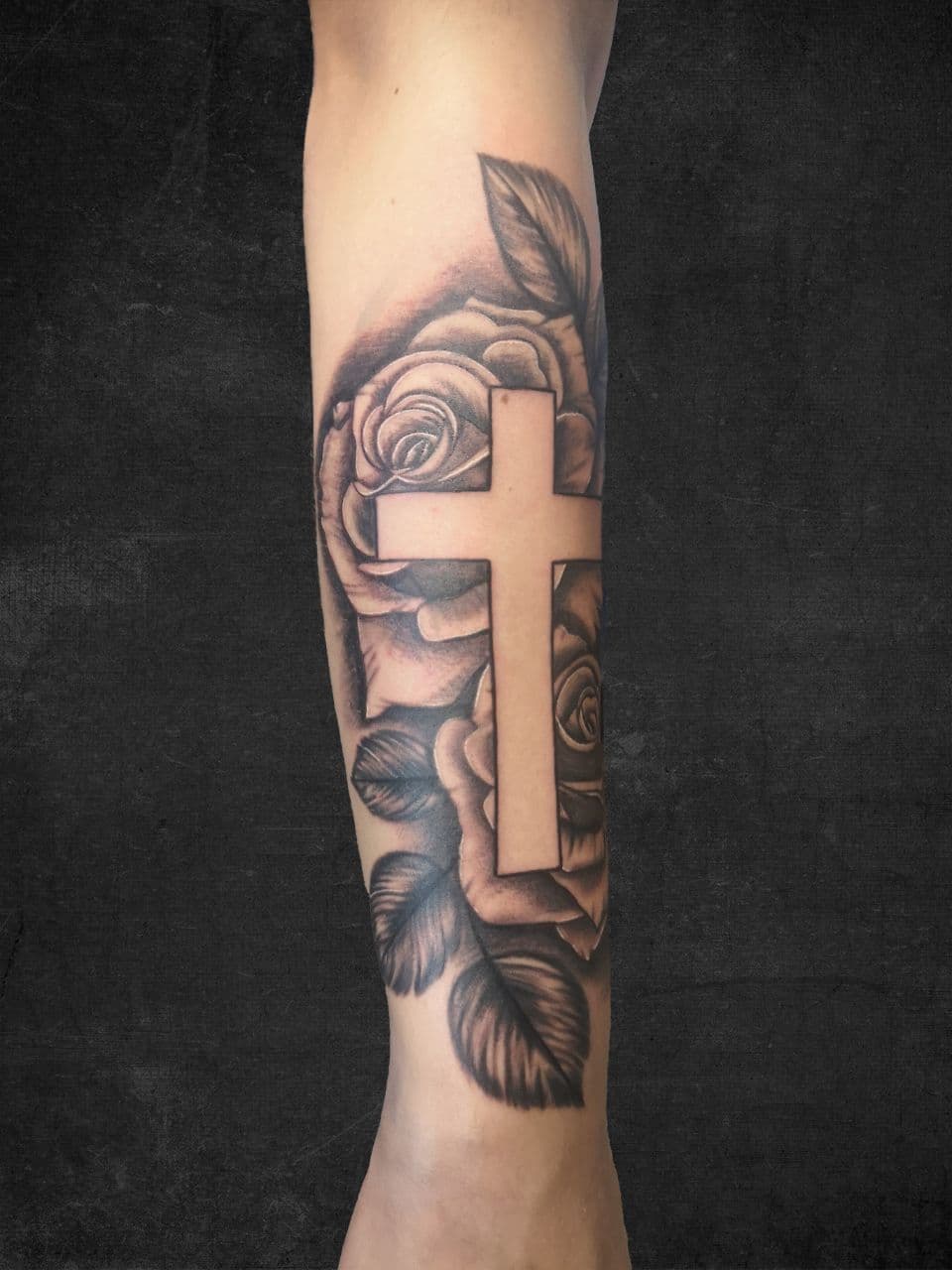Zwart-wit tattoo van een kruis omringd door rozen op de onderarm.