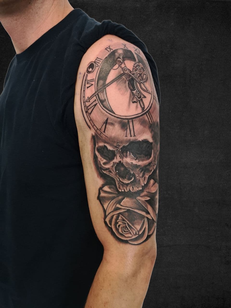 Realistische tattoo van klok, schedel en roos op bovenarm.