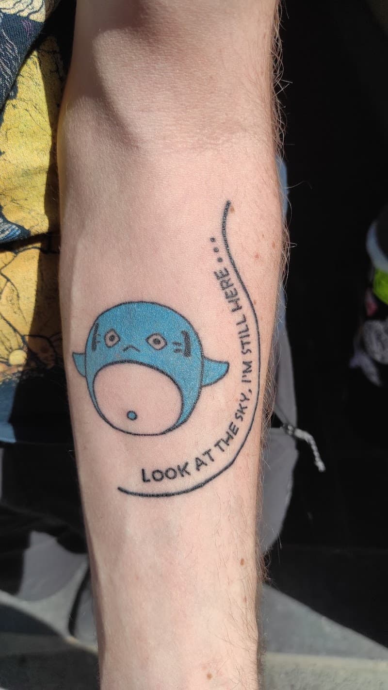 Blauw cartoon wezentje met tekst 'LOOK AT THE SKY, I'M STILL HERE...' op onderarm.