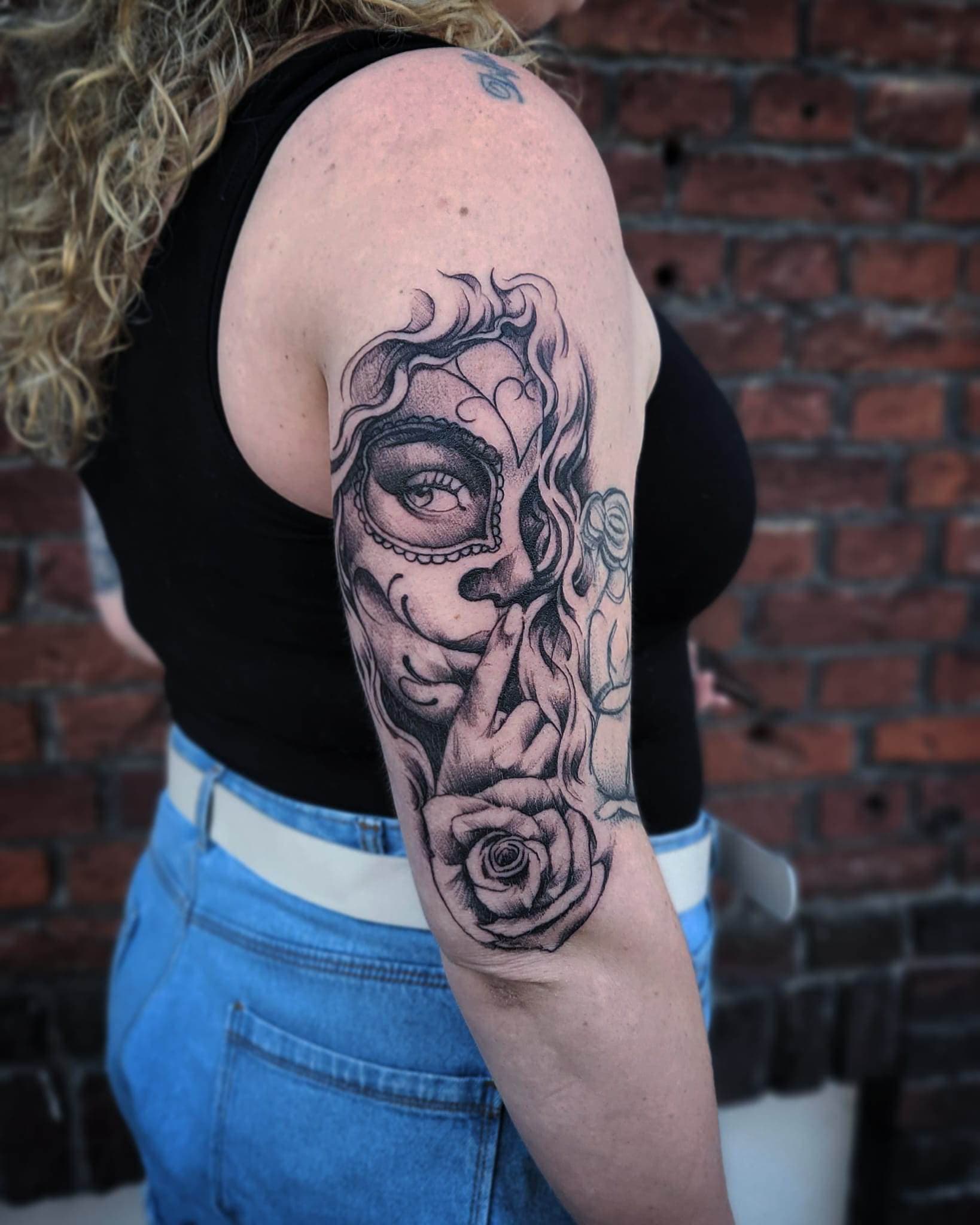 Zwart-wit tattoo van een vrouw met een doodskopmasker en een roos op de bovenarm.