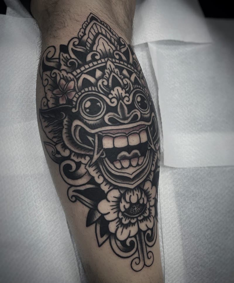 Zwart-wit Barong masker tattoo met bloemen op onderbeen.