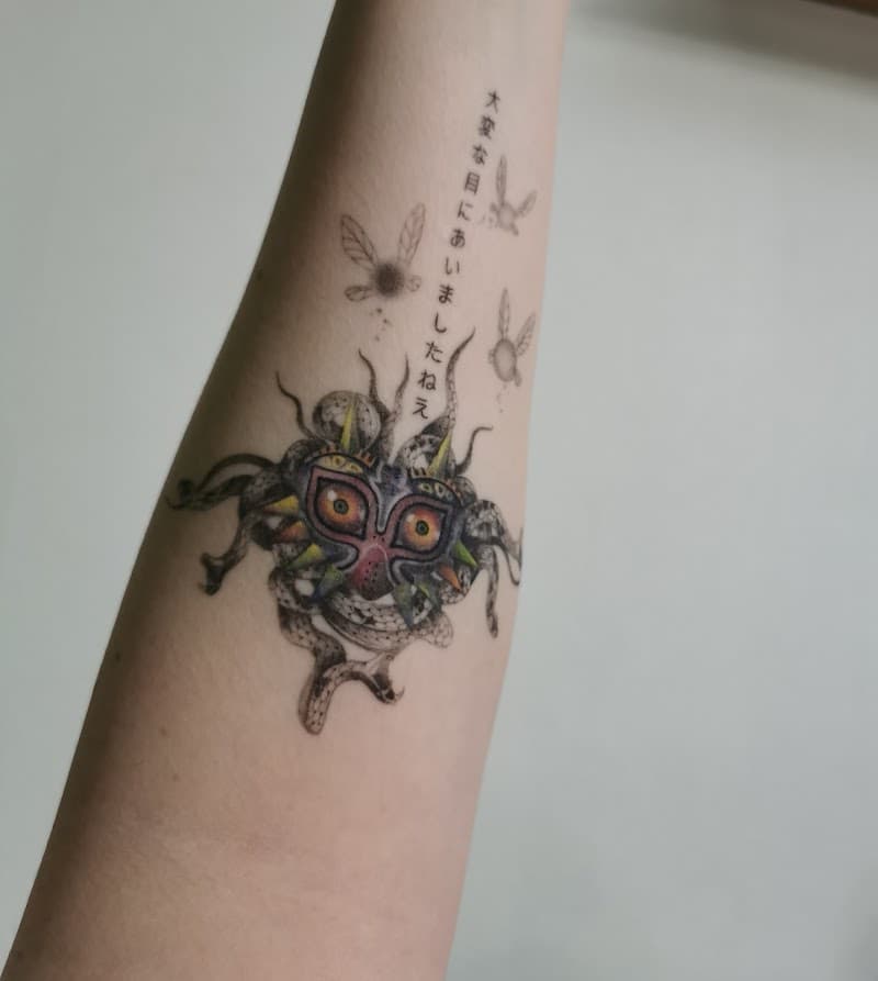 Illustratieve Majora's Mask tattoo met Japanse tekst en feeën op onderarm.