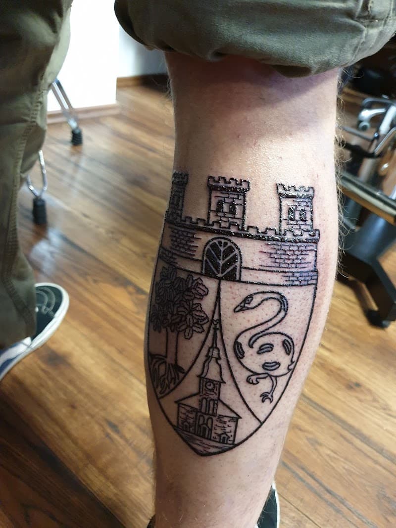Zwart-wit wapenschild tattoo met kasteel, zwaan, boom en kerk op kuit.