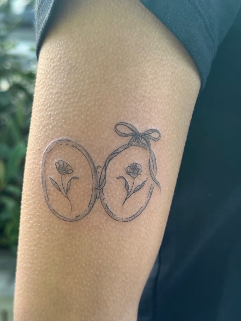 Fineline tattoo van twee medaillons met bloemen en een strik op de bovenarm.