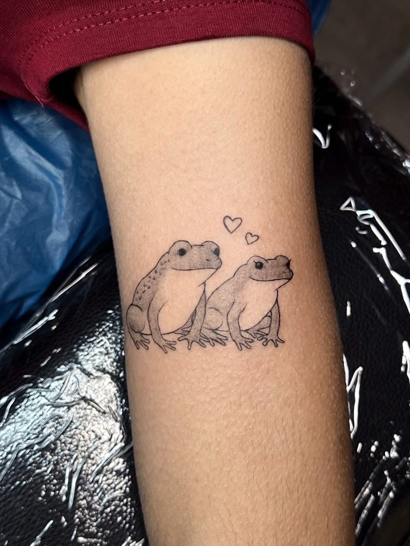 Schattige fineline tattoo van twee kikkers met hartjes op de bovenarm.