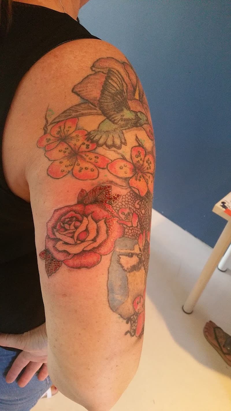 Kleurrijke tattoo met kolibrie, roos, kersenbloesem en pimpelmees op bovenarm.