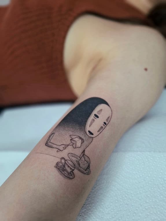 Illustratieve No-Face (Spirited Away) tattoo die taart eet op bovenarm.