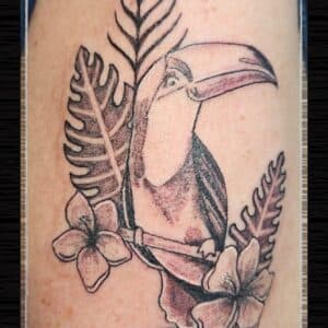 Zwart-wit tattoo van een toekan met tropische bladeren en bloemen.