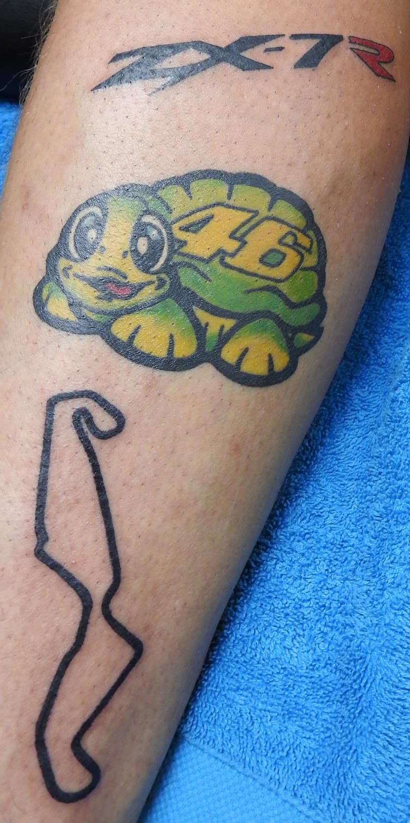 Drie tattoos op onderbeen: ZX-7R logo, Rossi's schildpad met 46, en een racecircuit.