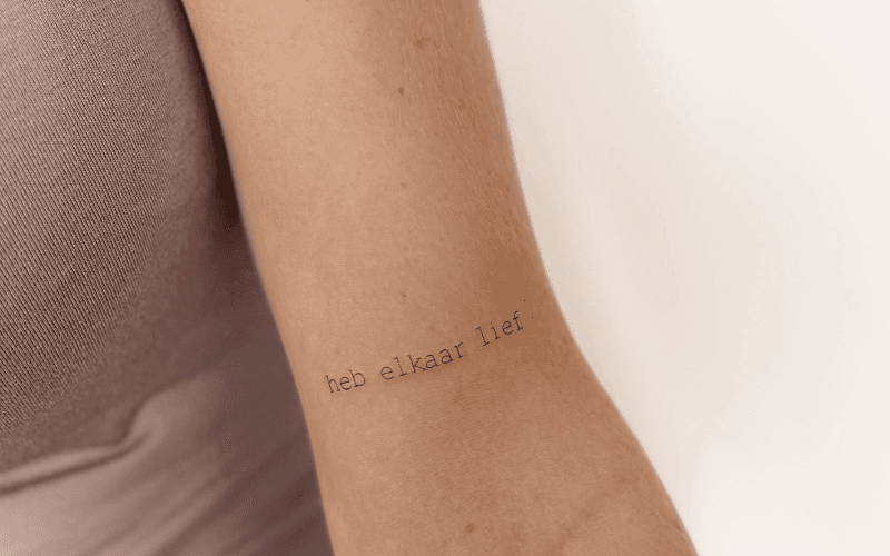Minimalistische 'heb elkaar lief' tekst tattoo op de binnenarm.