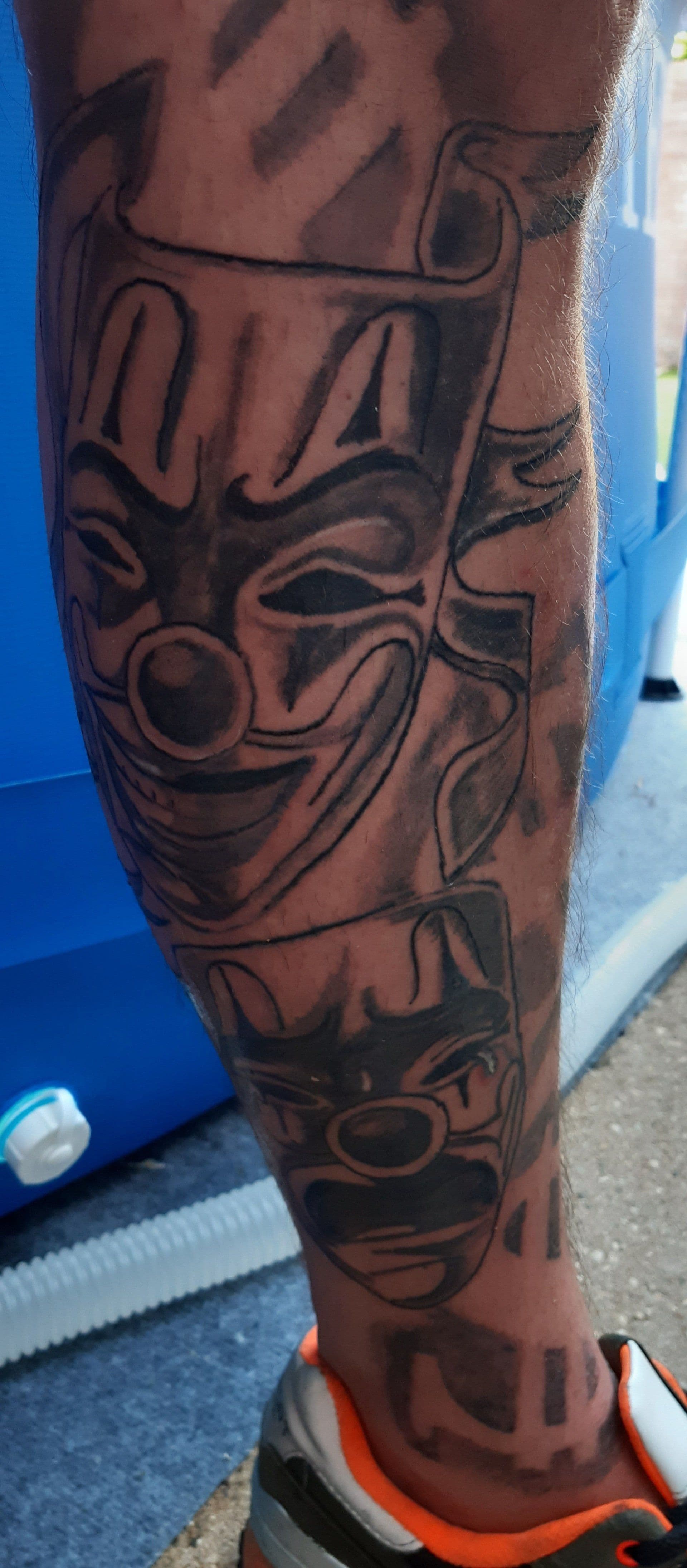 Zwart-wit tattoo van twee clown maskers op het onderbeen.