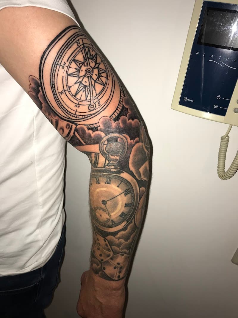 Zwart-wit tattoo van kompas, zakhorloge en dobbelstenen op arm.