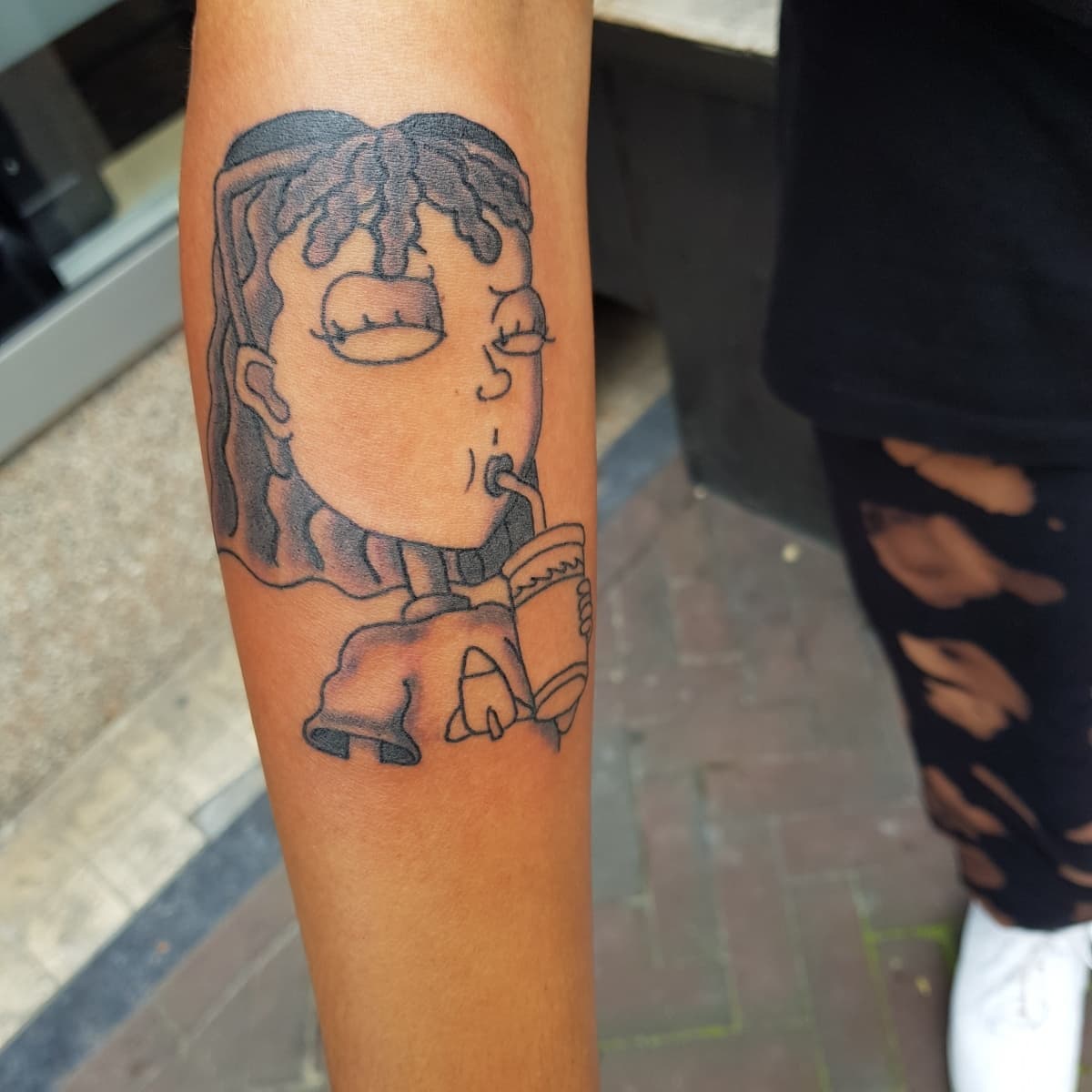 Blackwork cartoon karakter tattoo die uit een beker drinkt op de onderarm.